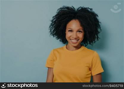 Happy dark-skinned woman, curly hair, smiles at camera, pleased expression. African female in yellow shirt. Blue background