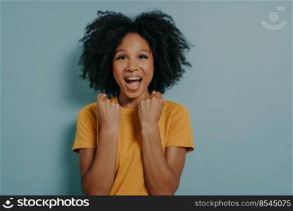 Happy dark skinned woman enjoys wonderful life, clenches fists with joy and screams. Young girl triumphs over success, feels excited. African lady rejoices goal achievement. Body language concept. Happy dark skinned woman enjoys wonderful life, clenches fists with joy and screams