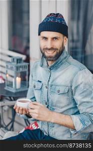 Happy delighted bearded middle aged male with positive expression, wears stylish hat and denim shirt, drinks hot tea, being in good mood, enjoys spare time. People, leisure and recreation concept