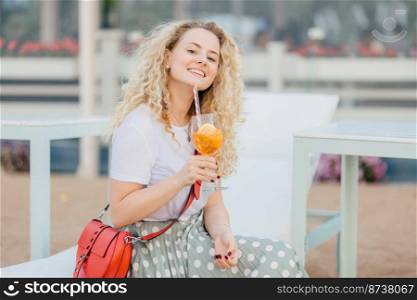 Happy delighted curly young woman dressed in white t shirt and long skirt, holds glass of cocktail, enjoys sunny day and good vacation, has manicure, smiles happily. People and vacation concept