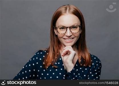 Happy delighted young woman keeps one hand under chin, smiles happily, wears transparent glasses, elegant clohtes, has eyes full of happiness, isolated over white background. Emotions concept
