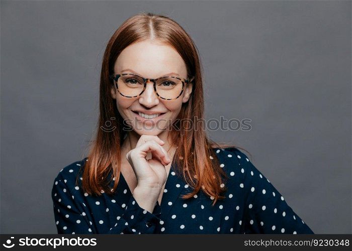 Happy delighted young woman keeps one hand under chin, smiles happily, wears transparent glasses, elegant clohtes, has eyes full of happiness, isolated over white background. Emotions concept