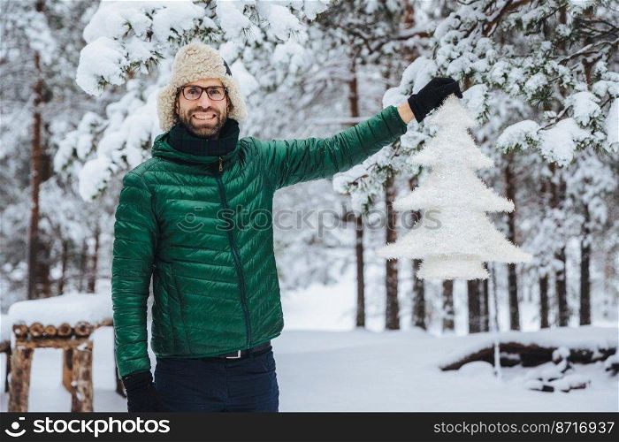 Happy delightful middle aged male with beard and mustache, wears warm jacket and hat, holds artificial white fir tree, spends morning in beautiful majestic forest. Weather and emotions concept