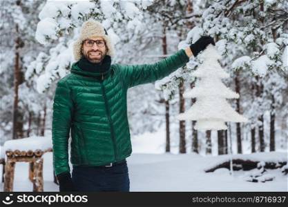 Happy delightful middle aged male with beard and mustache, wears warm jacket and hat, holds artificial white fir tree, spends morning in beautiful majestic forest. Weather and emotions concept