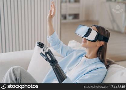 Happy disabled european girl with bionic cyber arm in virtual reality glasses gets treatment at home. Psychological rehabilitation for handicapped. Healthcare innovation healing technology.. Happy disabled european girl with bionic cyber arm in virtual reality glasses at home.