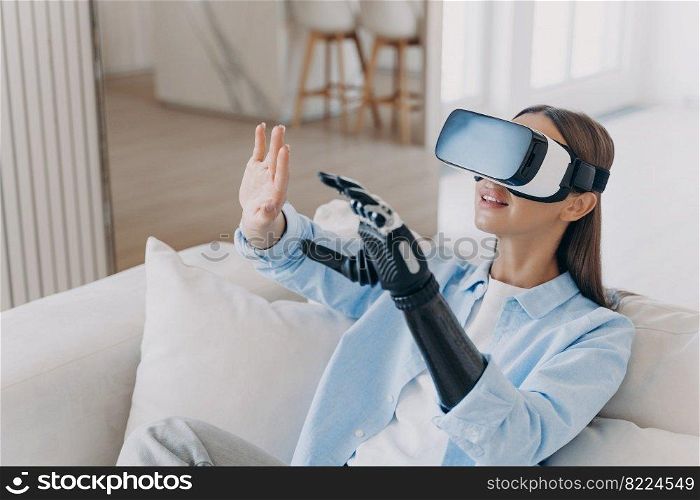Happy disabled girl wearing virtual reality glasses playing in video game, training to use her bionic prosthetic arm, sitting on sofa at home. High tech for rehabilitation of people with disabilities. Disabled girl in virtual reality glasses playing in video game, training to use prosthetic arm