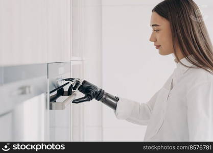 Happy disabled woman with robotic cyber hand is switching oven on. Amputee is cooking at home using bionic limb. Software in fingers and wrist joint. Innovation and orthopedic technologies.. Happy disabled woman with robotic cyber hand switching oven on. Cooking at home using bionic limb.