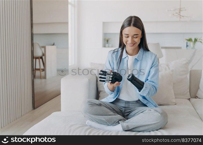 Happy disabled young woman is disassembling bionic arm prosthesis. Female&utee adjusting her artificial arm at home. European girl has myoelectric carbon hand. Electronic settings.. Happy disabled young woman is disassembling bionic arm prosthesis. Carbon myoelectric hand.