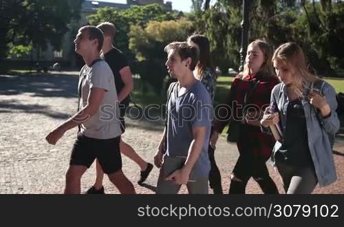 Happy diverse college students with backpacks and books walking through university campus to study. Cheerful group of teenage hipster friends going to lectures. Side view. Slow motion. Steadicam stabilized shot.