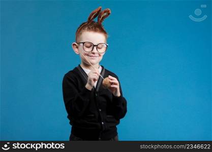 Happy Easter kids. Boy in rabbit bunny ears on head painting easter egg with paintbrush on blue background. Cheerful smiling child in glasses.. Happy Easter kids. Boy in rabbit bunny ears on head painting easter egg with paintbrush on blue background. Cheerful smiling child in glasses