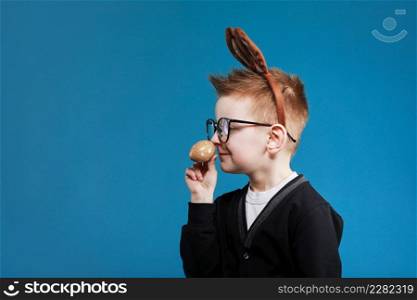 Happy Easter kids. Boy in rabbit bunny ears on head with colored egg on blue background. Cheerful smiling child in glasses.. Happy Easter kids. Boy in rabbit bunny ears on head with colored egg on blue background. Cheerful smiling child in glasses