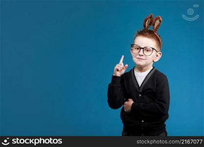 Happy Easter kids. child boy in rabbit bunny ears on head with glasses on blue studio background. Pointing up. Looking and smiling at camera. Happy Easter kids. child boy in rabbit bunny ears on head with glasses on blue studio background. Pointing up. Looking and smiling at camera.