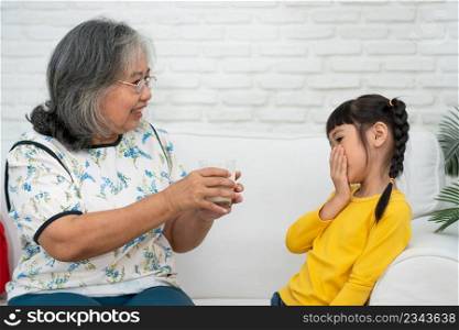 Happy elderly Asian Grandma sits beside her granddaughter and feeds fresh milk from glass for breakfast at home. Concept of a happy family and takes care together, preschool health care