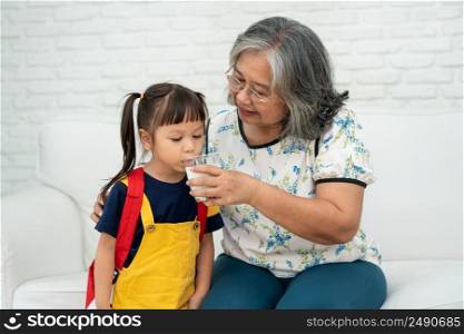 Happy elderly Asian Grandma sits beside her granddaughter and feeds fresh milk from glass for breakfast at home. Concept of a happy family and takes care together, preschool health care