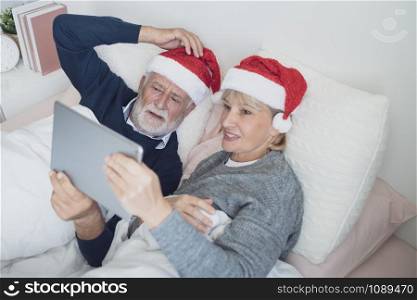 happy elderly couple caucasian senior man and woman with red hat talking family by using on tablet for christmas festival day in the morning in bedroom, retirement love family lifestyle concept