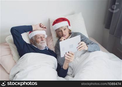 happy elderly couple caucasian senior man and woman with red hat talking family by using on tablet for christmas festival day in the morning in bedroom, retirement love family lifestyle concept