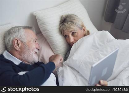 happy elderly couple caucasian senior man watching sport, game, TV, using on tablet and old woman sleep in white blanket in bedroom, retirement love lifestyle concept