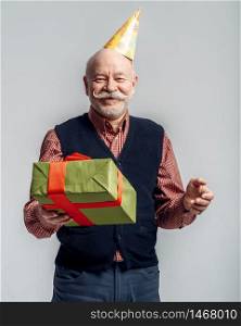 Happy elderly man in party cap holds gift box, grey background. Cheerful mature senior looking at camera in studio. Happy elderly man in party cap holds gift box