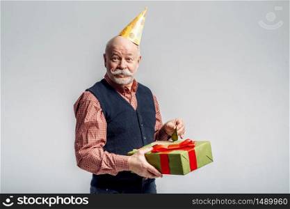 Happy elderly man in party cap holds gift box, grey background. Cheerful mature senior looking at camera in studio. Happy elderly man in party cap holds gift box