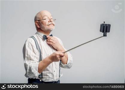 Happy elderly man makes selfie on phone, grey background. Mature senior showing his tongues at camera in studio. Happy elderly man makes selfie on phone