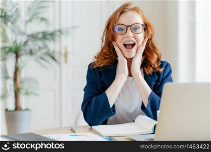 Happy emotive female office worker looks gladfully at camera, rejoices promotion and receiving money reward for good work, has ginger hair, weras spectacles, works on modern electronic notebook.