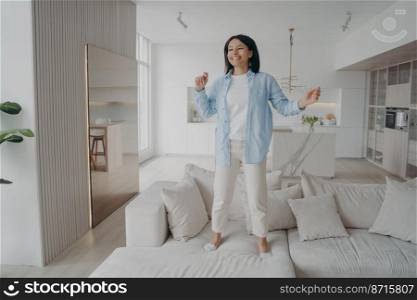 Happy energetic woman having fun, jumping on comfortable sofa alone at home, celebrating personal success. Joyful young female enjoying music, dancing on cozy couch in living room on weekend.. Happy energetic woman having fun, jumping on comfortable sofa alone at home, celebrating success