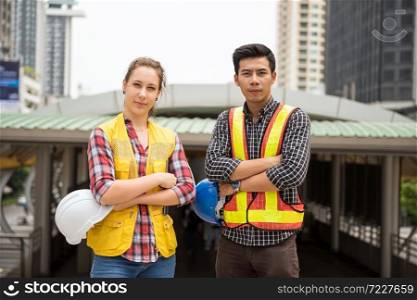 Happy Engineer team of Asian man and beauty American woman with safety helmet working on construction project in city. Teamwork for real estate high-rise site.