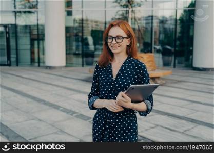 Happy European female entrepreeur with red hair wears spectacles and polka dot dress holds modern laptop looks positively into distance poses near office building outdoor glad after job interview.. European female entrepreeur with red hair wears spectacles and polka dot dress holds modern laptop