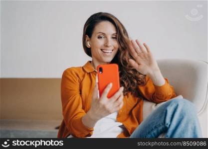 Happy european girl has video call at home. Cheerful young woman is using airpods and smartphone. Wireless earphones using at online conference. Technology using, leisure and relaxation concept.. Happy european girl has video call at home. Wireless earphones using at online conference.