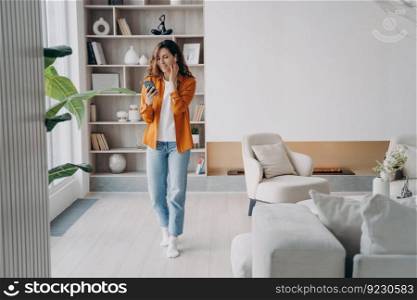 Happy european girl in headset has telephone call at home. Young woman is using airpods indoor. Wireless earphones. Relaxation, leisure and communication concept. Beautiful modern interior.. Happy european girl in headset has telephone call at home. Wireless earphones using.