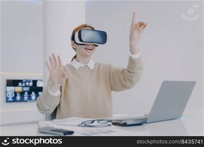Happy european girl in virtual reality goggles. Girl is sitting at the desk in modern office. Freelancer working on visual effects project in cyberspace. Concept of digital solutions.. Happy european girl in virtual reality goggles. Freelancer working on visual effects project.