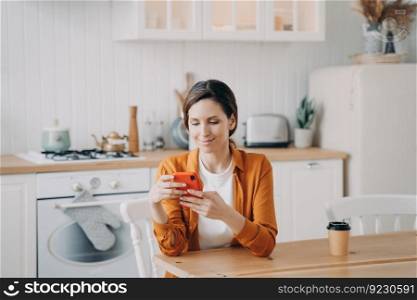 Happy european girl is sitting at the table at kitchen and texting on phone. Young woman in orange shirt is remote worker. Lady has coffee and browsing through internet. Workplace at home.. Happy girl is sitting at the table at kitchen and texting on phone. Workplace at home.