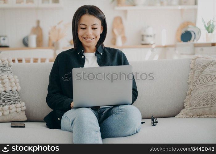 Happy european girl typing on computer. Business woman is working on startup project from home. Lady freelancer is sitting in comfortable couch with laptop and smiling, looking at the screen.. Happy girl typing on computer and smiling. Freelancer is sitting in comfortable couch with laptop.