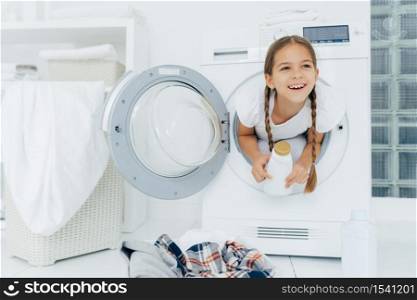 Happy European girl with two plaits, poses inside of wash machine, holds white bottle with liquid powder, has thoughtful expression, pile of dirty clothes in basin, looks somewhere into distance