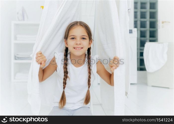 Happy European girl with two plaits, poses near clothes horse between white drying linen, poses in washing room against blurred background, white color. Children, cleanliness, washing concept