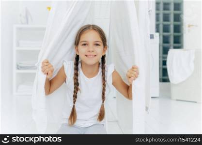 Happy European girl with two plaits, poses near clothes horse between white drying linen, poses in washing room against blurred background, white color. Children, cleanliness, washing concept