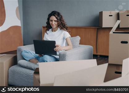 Happy european lady is sitting in armchair and working with pc. Delivery service ordering through internet. Mover among cardboard boxes is using laptop and smiling. Easy move concept.. Happy lady is sitting in armchair and working with pc. Delivery service ordering through internet.