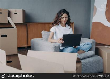 Happy european lady is sitting in armchair and working with pc. Delivery service ordering through internet. Mover among cardboard boxes is using laptop and smiling. Easy move concept.. Happy lady is sitting in armchair and working with pc. Delivery service ordering through internet.