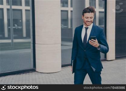 Happy european male entrepreneur in suit holding mobile phone and smiling, reading good news from business partner while standing outside on city street with laptop in hand, chatting with friend. Happy european male entrepreneur in suit holding mobile phone and smiling outdoors