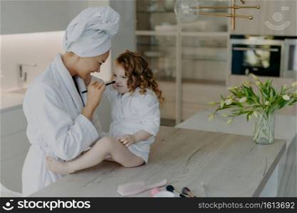 Happy European mother keeps cosmetic brush on daughters nose applies face powder dressed in white soft bath robe take care about appearance and skin. Family beauty wellbeing childhood motherhood