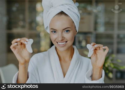 Happy European woman holds hydrogel patches in hands smiles toothily has healthy fresh skin looks positively at camera uses cosmetic product to remove dark circles under eyes wears bathrobe.