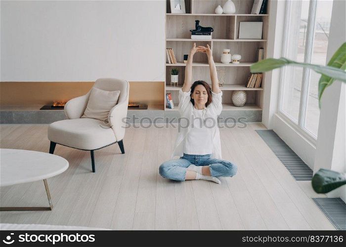 Happy european woman is meditating with her eyes closed. Girl is doing stretching exercises on floor at home. Lady sitting in lotus asana. Morning gymnastics. Concentration and balance concept.. Girl is doing stretching exercises on floor at home. Happy woman is meditating with her eyes closed.