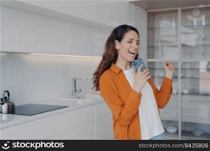 Happy european woman is singing with phone as with microphone. Girl having fun in her living room. Application for music listening online. Favourite song singing at home, relaxation and dancing.. Happy european woman is singing with phone as with microphone. Favourite song singing at home.