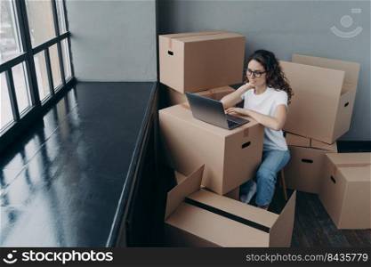 Happy european woman sitting among cardboard boxes. Lady is in waiting for shipping service near window. Girl with laptop on box. Relocation to new apartment.. Happy european woman sitting among cardboard boxes waiting for shipping service near window.