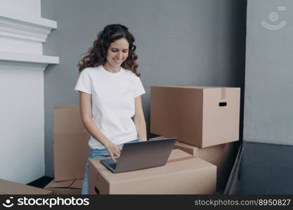 Happy european woman unloading cardboard boxes at new place. Girl in white t-shirt is typing on laptop on the box. Relocation and distance work concept.. Happy european woman unloading cardboard boxes at new place and typing on laptop on the box.