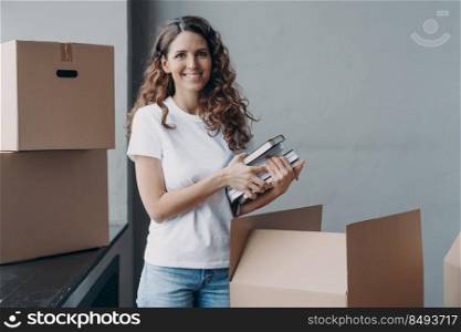 Happy european woman unpacking boxes and taking off books and notes. Young student is housing in campus room. Girl in white t-shirt indoors. Relocation and independence concept.. Happy european woman unpacking boxes and taking off books and notes. Young student in campus room.