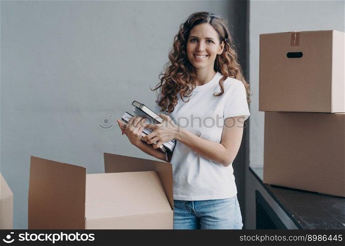 Happy european woman unpacking boxes and taking off books and notes. Young student is housing in c&us room. Girl in white t-shirt indoors. Relocation and independence concept.. Happy european woman unpacking boxes and taking off books and notes. Young student in c&us room.