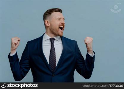 Happy excited businessman in suit celebrating success in business startup and keeping arms raised while standing against light steel blue background, male employee got job promotion. Excited businessman in suit keeping arms raised and screaming while celebrating success