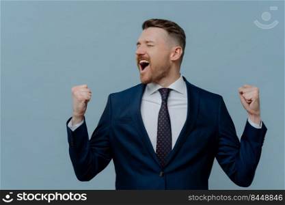 Happy excited businessman in suit celebrating success in business startup and keeping arms raised while standing against light steel blue background, male employee got job promotion. Excited businessman in suit keeping arms raised and screaming while celebrating success