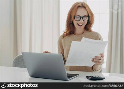 Happy excited redhead woman reading paper letter with notification about last mortgage payment while calculating family monthly budget and paying bills online at home, using laptop and calculator. Happy excited redhead woman reading paper letter with notification about last mortgage payment